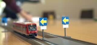 Auf dem Bild sind zwei Gleise einer Modelleisenbahn zu sehen. Auf dem linken Gleis fährt eine rote Regionalmodelleisenbahn. Vor dem Zug sind jeweils rechts der beiden Gleise zwei blaue Schilder mit einem gelben nach links zeigenden Pfeil darauf. Auf dem Bild sind zwei Gleise einer Modelleisenbahn zu sehen. Auf dem linken Gleis fährt eine rote Regionalmodelleisenbahn. Vor dem Zug sind jeweils rechts der beiden Gleise zwei blaue Schilder mit einem gelben nach links zeigenden Pfeil darauf.