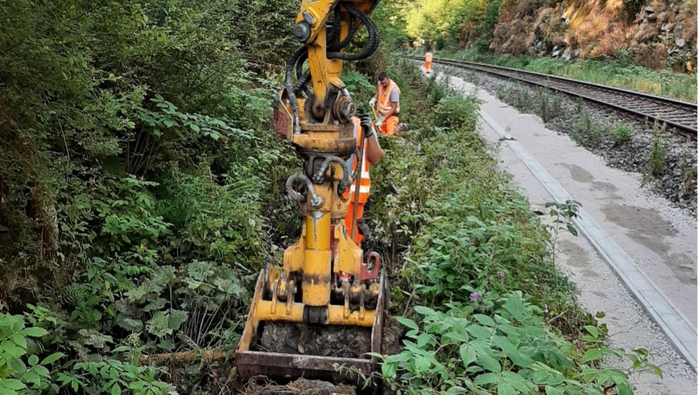 Zu erkennen ist ein Baggerschaufel neben einem Gleisfeld. Drei Menschen in Warnweste stehen im unmittelbaren Umfeld und beobachten die Bauarbeiten.
