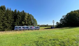 blauer Testzug fährt im Digitalen Testfeld Bahn auf einem Gleis. Hinter dem Zug sind Tannen zu sehen, im Vordergrund eine Wiese. In der rechten Bildhälfte ist ein Funkmast | © DB InfraGO AG blauer Testzug fährt im Digitalen Testfeld Bahn auf einem Gleis. Hinter dem Zug sind Tannen zu sehen, im Vordergrund eine Wiese. In der rechten Bildhälfte ist ein Funkmast | © DB InfraGO AG