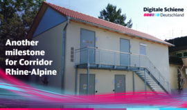 You can see a bright building with four steel doors and a steel staircase on the façade leading to the second floor. The sign in front of it reads: “Another milestone for the Rhine-Alpine corridor” You can see a bright building with four steel doors and a steel staircase on the façade leading to the second floor. The sign in front of it reads: “Another milestone for the Rhine-Alpine corridor”