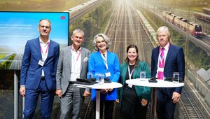 usanne Henckel, Staatssekretärin im Bundesministerium für Digitales und Verkehr, Axel Schuppe, Geschäftsführer des Verbandes der Bahnindustrie Deutschland e.V., Jan Görnemann, Sprecher der Geschäftsführung des Bundesverbandes SchienenNahverkehr e.V. und Dr. Volker Hentschel, Vorstandsbeauftragter Digitale Schiene Deutschland bei DB InfraGO AG stehen zwischen zwei Stehtischen und einem Monitor. Im Hintergrund ist eine Fotowand, auf der mehrere Gleise mit Zügen zu sehen sind.