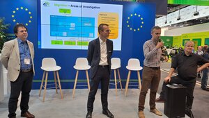 Drei Männer stehen an einem Messestand und halten einen Vortrag. Im Hintergrund ist eine Messewand in blau mit Logo der EU sowie ein Bildschirm mit einer Präsentation