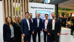 Zwei Frauen und fünf Männer stehen am Messestand der Digitalen Schiene Deutschland. Im Hintergund ist ein Raumtrenner aus Holz sowie eine weiße Wand mit einem Bildschirm. Über dem Bildschirm steht "Digitale Schiene Deutschland"
