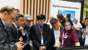 Mehrere Menschen stehen vor einem Bildschirm am Messestand. Eine Frau zeigt mit dem Finger auf den Bildschirm 