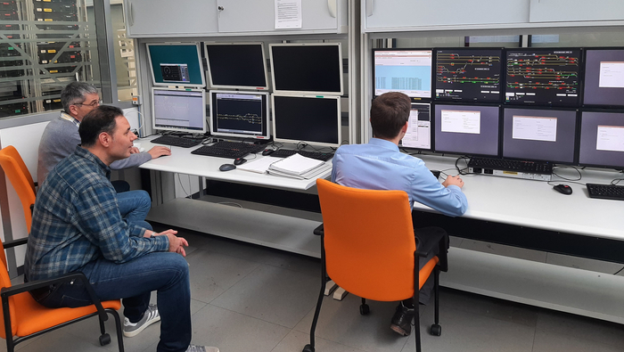 Alexander Gehrecke, Firas Omran und Alexander Schuler sitzen vor zwei Schreibtischen mit 14 Monitoren während der Abnahme.