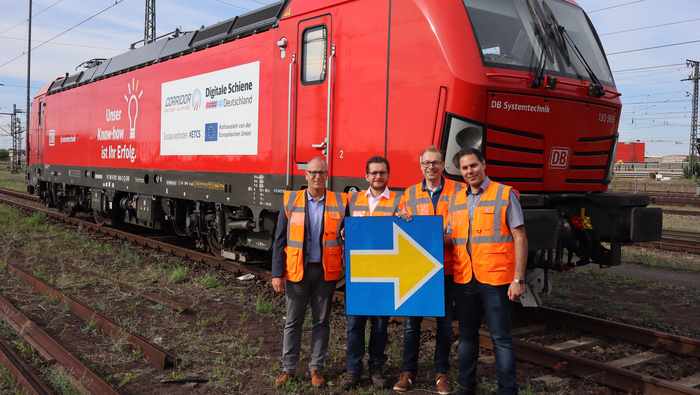Ulrich Kohlenberger, Frederik Schmalle, Marc Hansen und Firas Omran stehen im Gleisfeld vor einer roten Lokomotive.
