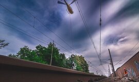 Ein Bild der die Strommasten der Eisenbahnstrecke zeigt und einen Helikopter im himmel zeigt.
