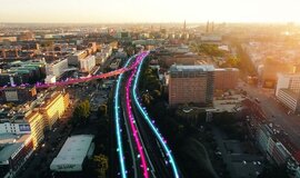 Stadtsicht im Sonnenaufgang von oben mit zweigleisiger Bahnstrecke, in die von links zwischen Häuserreihen ein weiteres Gleis einfädelt. Auf den Gleisen sind pinke und neonblaue Linien, auf denen kleine Hashtags schweben. Stadtsicht im Sonnenaufgang von oben mit zweigleisiger Bahnstrecke, in die von links zwischen Häuserreihen ein weiteres Gleis einfädelt. Auf den Gleisen sind pinke und neonblaue Linien, auf denen kleine Hashtags schweben.