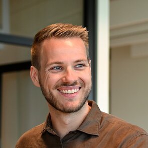 Protraitaufnahme von Felix Grob: Ein lächelnder Mann mit blonden kurzen Haaren und Jeansfarbenem Hemd