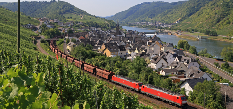 Zu sehen ist eine Ortschaft im Grünen, an der rechts ein Fluss vorbeifährt. Links der Ortschaft führ eine eingleisige Strecke, auf der ein Güterzug mit zwei roten Triebfahrzeugen fährt. Zu sehen ist eine Ortschaft im Grünen, an der rechts ein Fluss vorbeifährt. Links der Ortschaft führ eine eingleisige Strecke, auf der ein Güterzug mit zwei roten Triebfahrzeugen fährt.