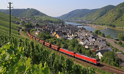 Ein Güterzug fährt durch die Landschaft Ein Panoramablick zeigt ein roter Zug, diese zieht mehrere Güterwagons hinter sich. Im Vordergrund ist ein Hang mit Weinreben. Hinter dem Zug befindet sich eine kleine Stadt. Hinter der Stadt ist der Rhein zu sehen umgeben von mehreren kleinen Bergen.