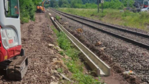 A small excavator in the foreground and a larger one in the background are working on railway tracks. There are mountains and green trees in the background.