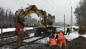 A yellow excavator is positioned on the tracks and is working on the track in front of it. Two people in high-visibility waistcoats are working on the ground, while a third person in a high-visibility waistcoat oversees the process.