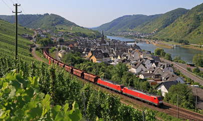 A panoramic view shows a red train pulling several cargo wagons behind it. In the foreground is a slope with vines. Behind the train is a small town. The Rhine can be seen behind the town, surrounded by several small mountains.