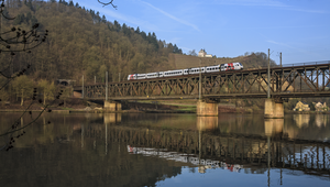 A train is travelling over an old iron bridge. Which is located over a river and a mountain with a forest can be seen in the background. The trees no longer have any leaves. There is also a small fitter and a vineyard on the mountain.