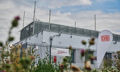 Digitales Stellwerk Meitingen - Mertingen Es ist eine Seitenansicht von einem weißen Gebäude zu sehen, im Vordergrund befinden sich grüne Pflanzen und eine Weiße Flagge mit rotem DB Logo.