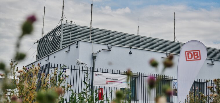 A side view of a white building is shown, with green plants and a white flag with a red DB logo in the foreground.