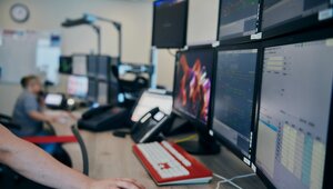 A workstation with six screens can be seen. A hand on the workstation is operating a computer mouse. In the background another workstation with many screens where a person is working can be seen.