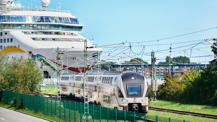 Das Foto zeigt einen weißen Doppelstock-Intercity. Neben den Gleisen befindet sich ein grüner Zaun, über den Gleisen Oberleitungen. Im hinteren Abschnitt des Bildes ist ein Kreuzfahrtschiff und deren Brücke zu erkennen. Das Foto zeigt einen weißen Doppelstock-Intercity. Neben den Gleisen befindet sich ein grüner Zaun, über den Gleisen Oberleitungen. Im hinteren Abschnitt des Bildes ist ein Kreuzfahrtschiff und deren Brücke zu erkennen.