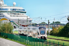 Das Foto zeigt einen weißen Intercity-Zug. Im Hintergrund ist ein Kreuzfahrtschiff zu erkennen.