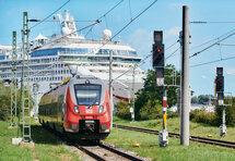 Das Foto zeigt einen roten Regionalzug. Im Hintergrund ist ein Kreuzfahrtschiff zu erkennen.