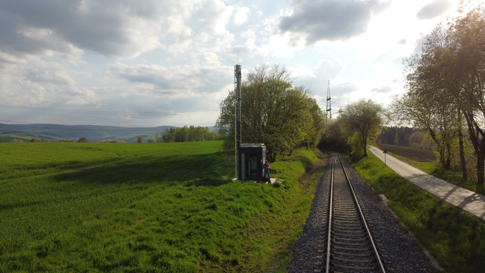 Foto zeigt eingleise Eisenbahnstrecke im Erzgebirge. Auf der Linken der Gleise ist ein FRMCS/5G Funkmast zu sehen. Auf der linken Bildhälfte befindet sich ein grünes Feld.