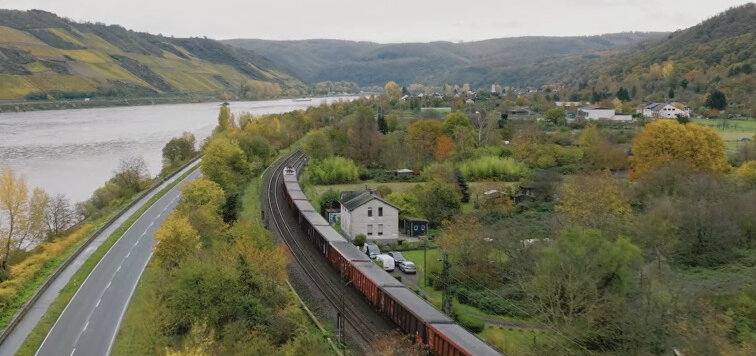 Von links nach Rechts: Ein Fluss, eine zweispurige Straße und eine zweigleisige Bahnstrecke  mit einem Güterzug. Daneben ein Haus.