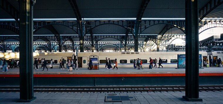 Ansicht eines Bahnhofs mit Blick auf den gegenüberliegenden Bahnsteig. Im Hintergrund steht ein beigefarbener Zug. Menschen laufen auf dem Bahnsteig. Ansicht eines Bahnhofs mit Blick auf den gegenüberliegenden Bahnsteig. Im Hintergrund steht ein beigefarbener Zug. Menschen laufen auf dem Bahnsteig.