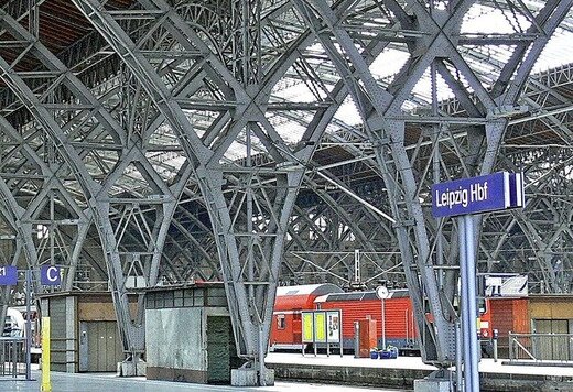 Bahnhofsgebäude mit Stahlträgern. Ein roter Regionalzug ist im Hintergrund zu sehen. Auf dem Bahnhofsschild ist "Leipzig HBF" zu lesen.