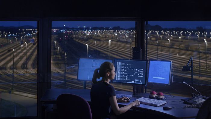 A Women sitting in front of a desk with 3 screens depicting the rail system. The Background consists of Big windows where a lot of traintracks are visible. The Scenario is shown during the nighttime.