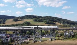 Ein ICE der über eine Brücke fährt. Unter der Brücke sind Häuser erkennbar und im Hintergrund sind grüne Hügel und Wälder.