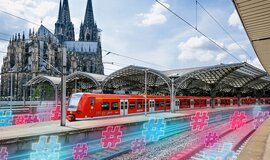 einer roter Regionalzug steht im Bahnhof Köln. Hinter dem Zug ist der Kölner Dom zu sehen. Neonfarbene Hashtags schweben auf den Gleisen.