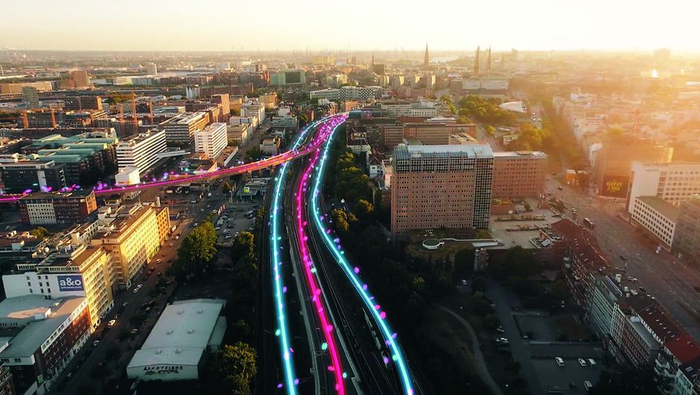 Luftaufnahme einer Großstadt. In der Mitte des Bildes befinden sich Gleise welche durch Blaue und Pinke Streifen hervorgehoben sind.